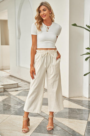 a woman standing on a tile floor wearing a white top and wide legged pants