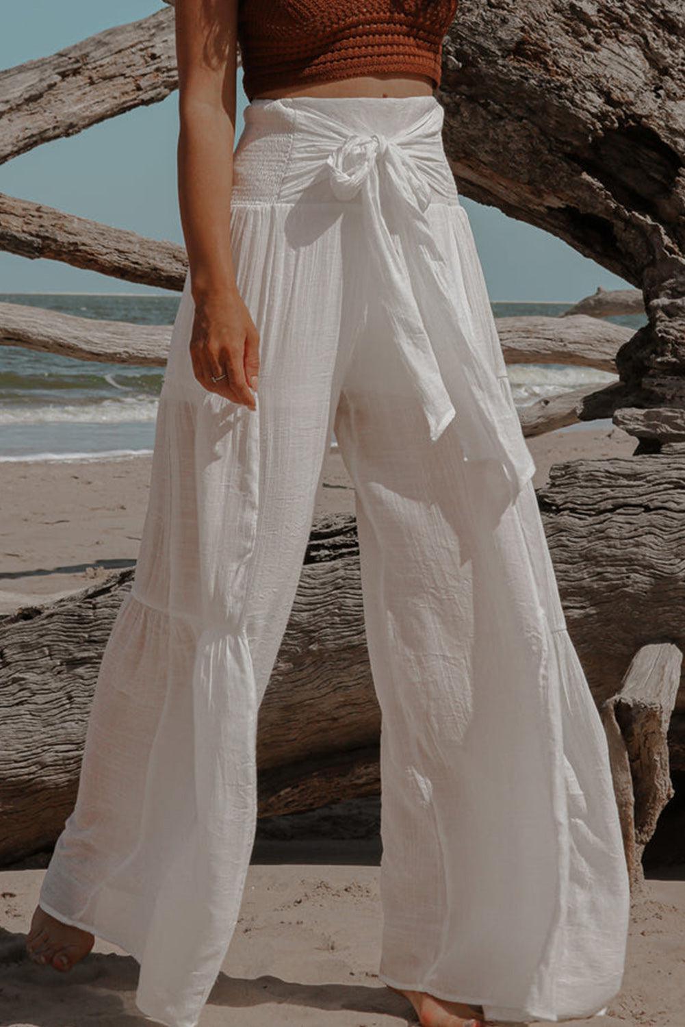 a woman standing on a beach wearing a brown top and white pants