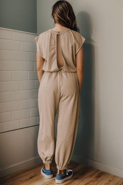 a woman standing against a wall wearing a tan jumpsuit